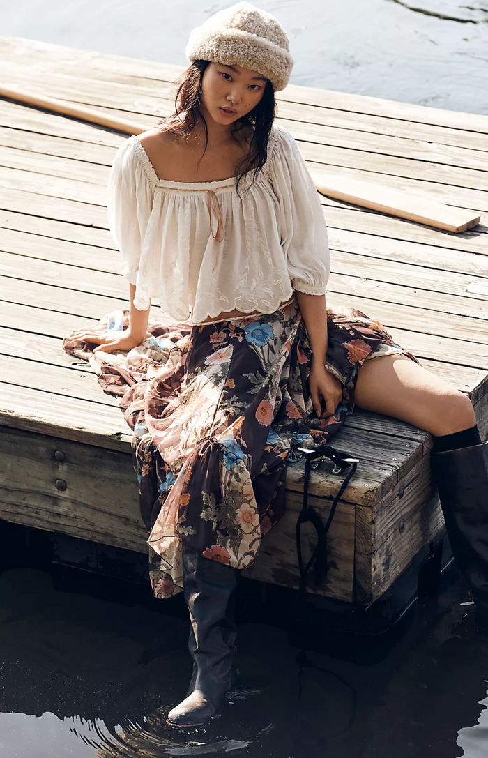 Woman sitting on a wooden dock wearing a floral skirt and pearl blouse.