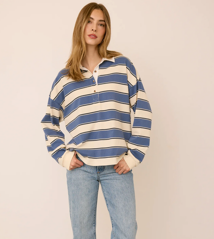 A woman modeling a blue and cream striped long-sleeve polo shirt with light-wash blue jeans, standing against a plain beige studio background.