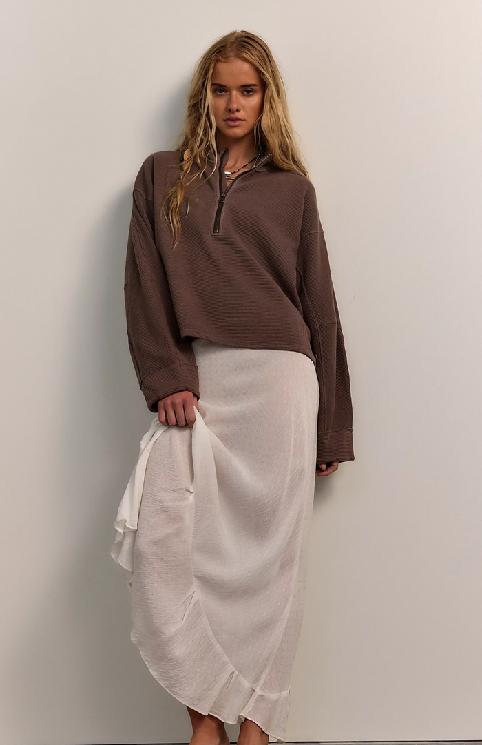Woman wearing a mocha quarter zip top and white skirt against a plain background