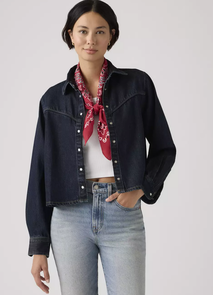 Woman wearing a dark denim jacket, white shirt, light blue jeans, and red bandana on a plain background