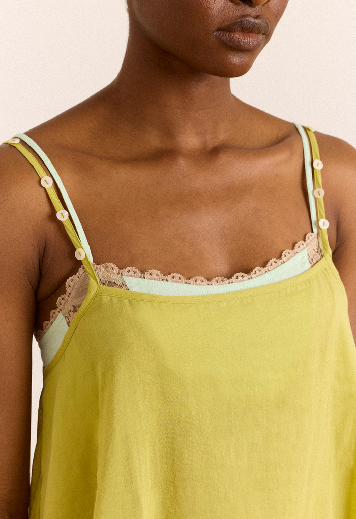 A close-up of a model wearing a chartreuse camisole top with thin straps, lace trim, and decorative buttons.