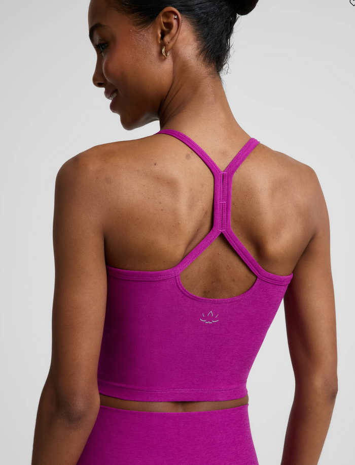 Back view of a woman wearing a berry bliss-colored sports bra with a crossover criss-cross back strap design and a small white logo, paired with matching high-waisted leggings against a light gray studio background.