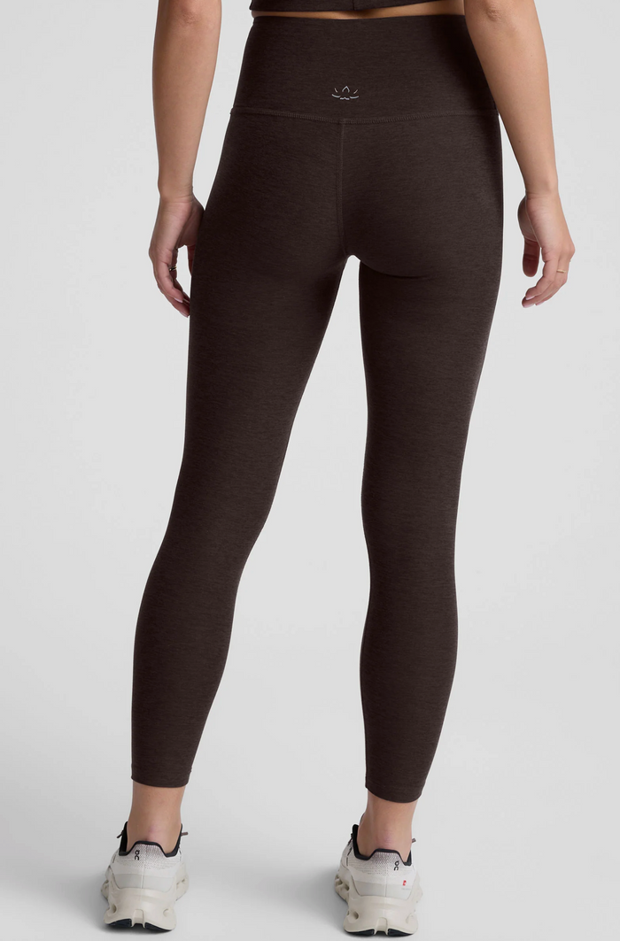A person shown from the back wearing chocolate-colored high-waisted leggings and white sneakers against a light gray studio background.
