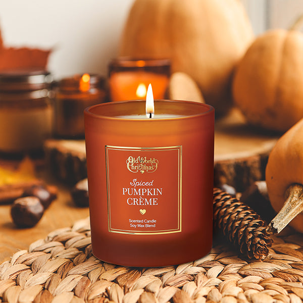 A lit orange candle labeled Spiced Pumpkin Crème sits on a woven mat beside pine cones and autumn decor, with pumpkins and blurred candles in the background. The scene feels warm and cozy.