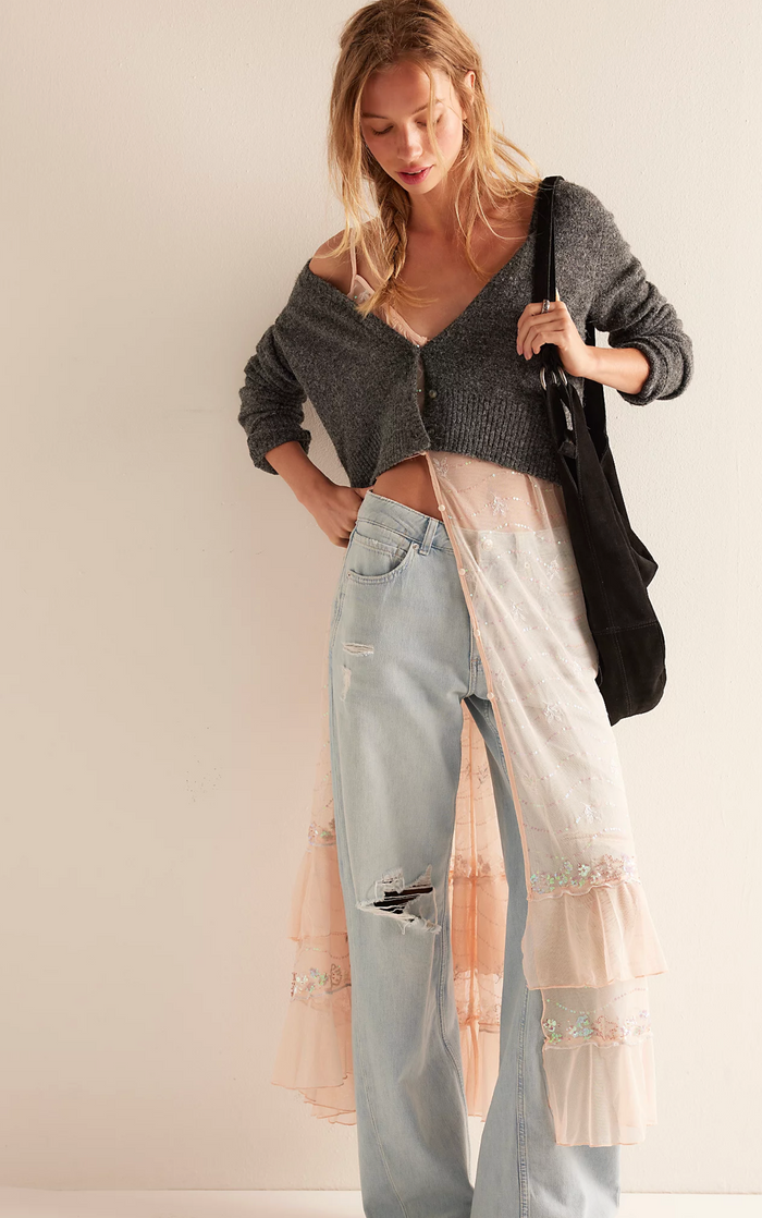 Woman wearing a gray cardigan over a sheer top with ripped jeans on a plain background