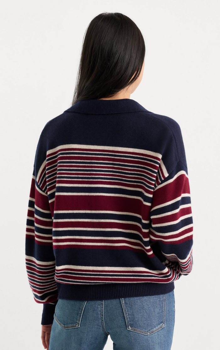 A woman viewed from behind wearing a navy blue knit sweater with horizontal burgundy and cream stripes across the body and sleeves, paired with blue jeans against a plain white studio background.