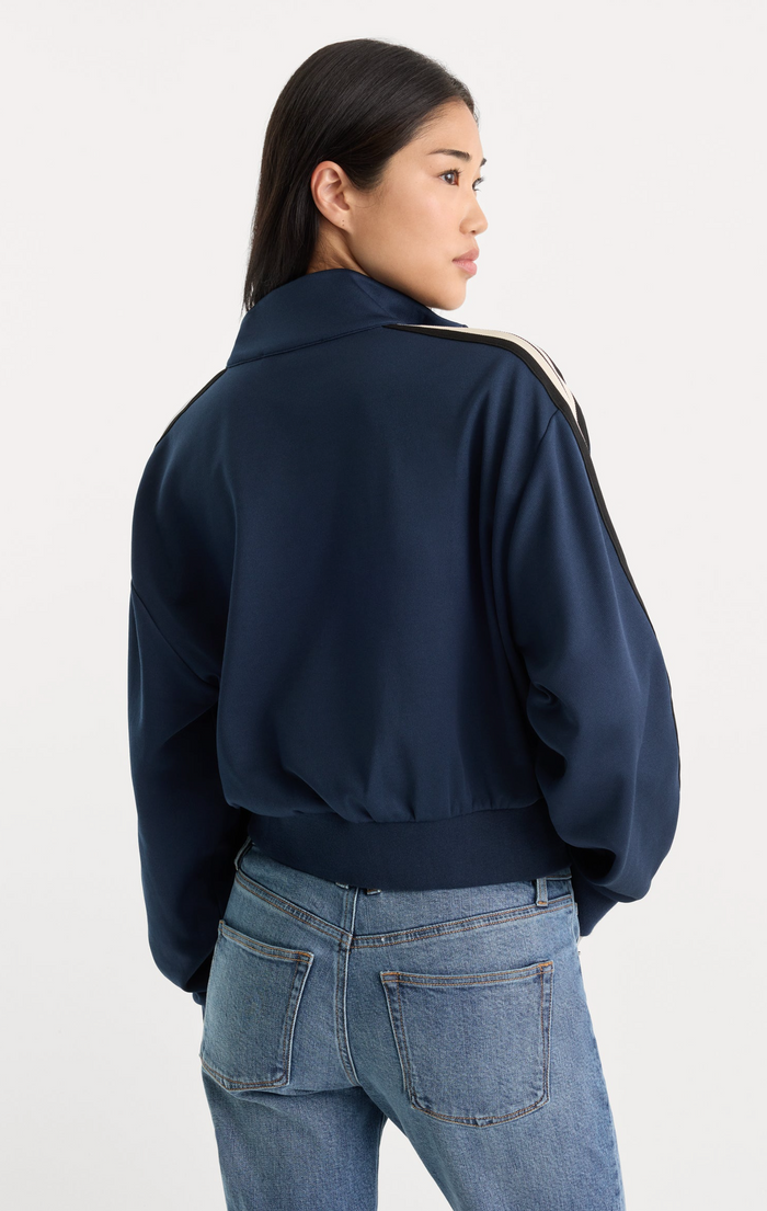 A woman shown from the back wearing a navy blue bomber jacket with gold stripes along the shoulders and blue jeans, against a plain light gray studio background.