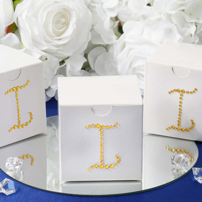 White gift boxes with jeweled initials on a mirrored surface surrounded by flowers.