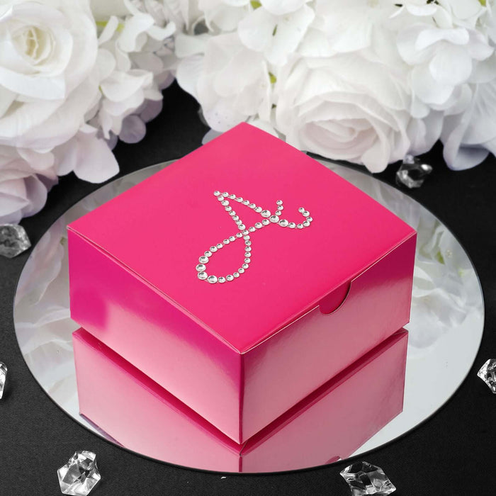 Pink gift box with jeweled letter "A" on a mirrored surface surrounded by white flowers.