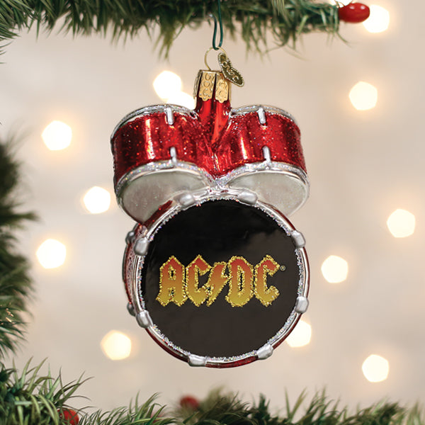 A Christmas tree ornament shaped like a red drum set with AC/DC written in yellow on the bass drum, hanging on a branch with blurred lights in the background.