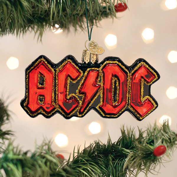 A Christmas tree ornament shaped like the AC/DC band logo, featuring bold red letters with a lightning bolt in the center, hanging from a green tree branch with blurred holiday lights in the background.