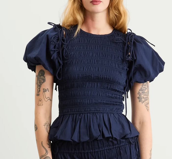 A woman wearing a navy blue smocked dress with puffed sleeves, tie details on the shoulders, and a ruffled waist, shown from the neck down against a light gray background.
