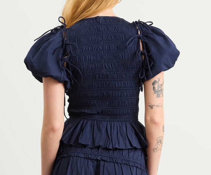 Back view of a navy blue smocked dress with puffed sleeves, tie details on the shoulders, and a ruffled waist, shown on a model against a light gray background.