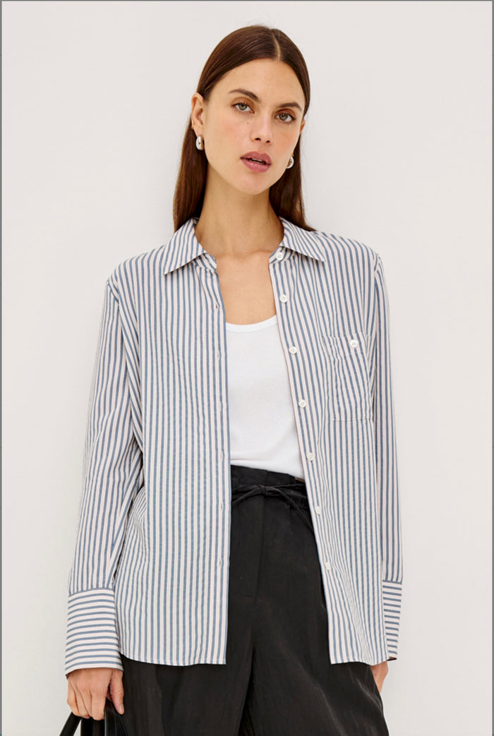 A woman modeling a long-sleeve button-up shirt with thin vertical navy and white stripes, worn open over a white tank top and black trousers, against a plain light white studio background.