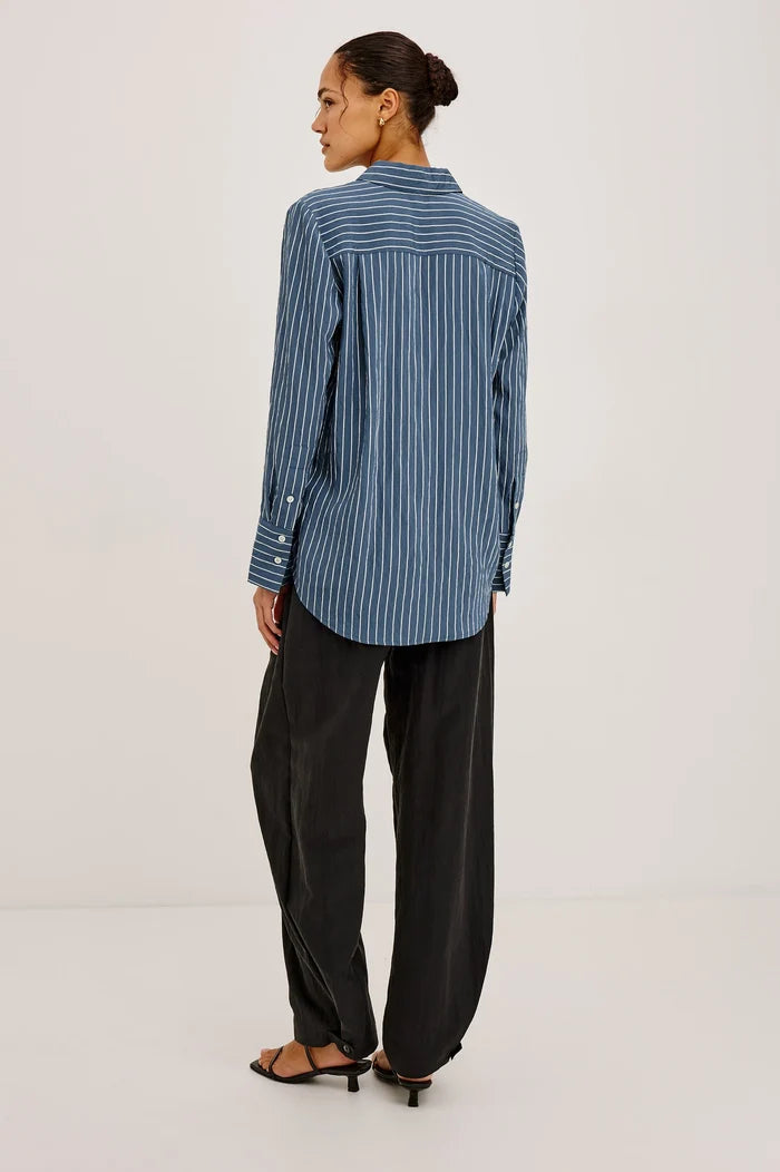 Woman with dark hair wearing a blue and white vertical striped button down with her back side facing and black trousers.