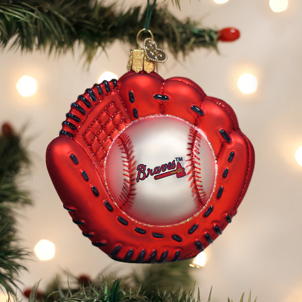 A Christmas tree ornament shaped like a red baseball glove holding a baseball with the Braves logo, hanging from a tree branch with blurred lights in the background.