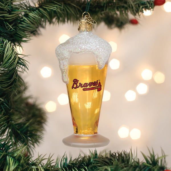 A Christmas tree ornament shaped like a beer glass with glittery foam, featuring the Braves logo, hanging on a green tree branch with blurred lights in the background.