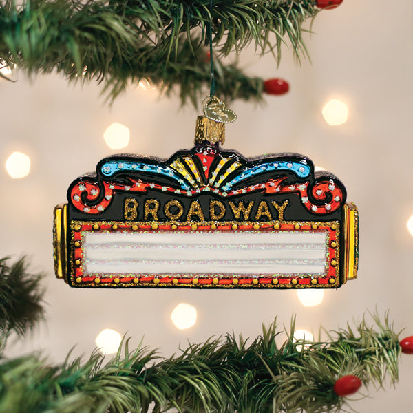 A colorful, glittery ornament shaped like a Broadway theater marquee hangs on a Christmas tree, with the word BROADWAY on top and blank space below, surrounded by green branches and soft, blurred lights in the background.