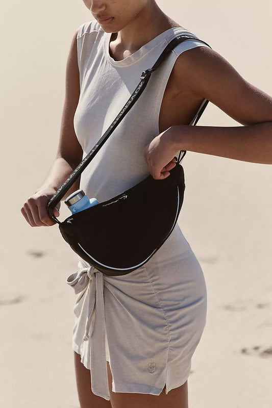 A person wearing a beige dress is carrying a black sling bag with reflective detailing and an adjustable shoulder strap.
