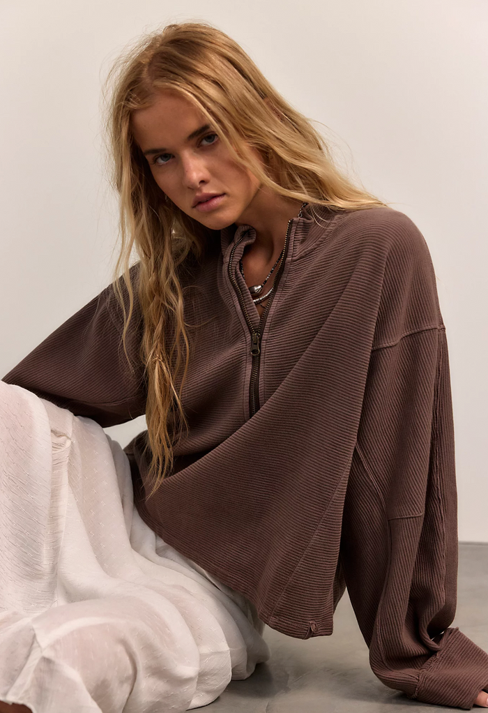 Woman wearing a mocha oversized quarter zip shirt sitting on the floor against a plain background