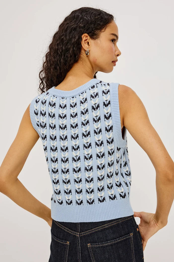 A woman shown from the back wearing a light blue sleeveless knit top with a repeating black and white butterfly pattern, paired with dark blue jeans against a light gray studio background.