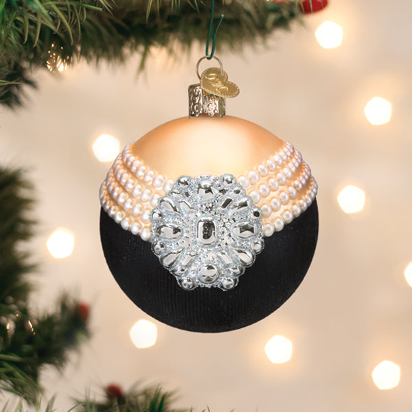 A Christmas tree ornament decorated to resemble a black evening gown with pearl necklace details and a jeweled brooch, hanging on a tree with blurred lights in the background.