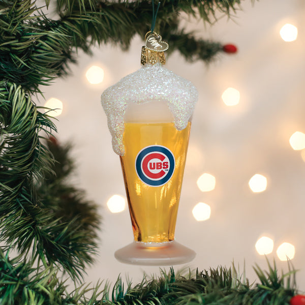 A glass ornament shaped like a beer glass with foam on top, featuring the Chicago Cubs logo, hangs on a Christmas tree with blurred lights in the background.