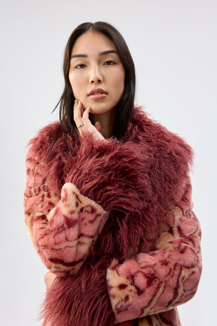 Close-up of a model wearing a pink and beige animal-print faux fur coat with a burgundy shaggy collar, hand resting near the face