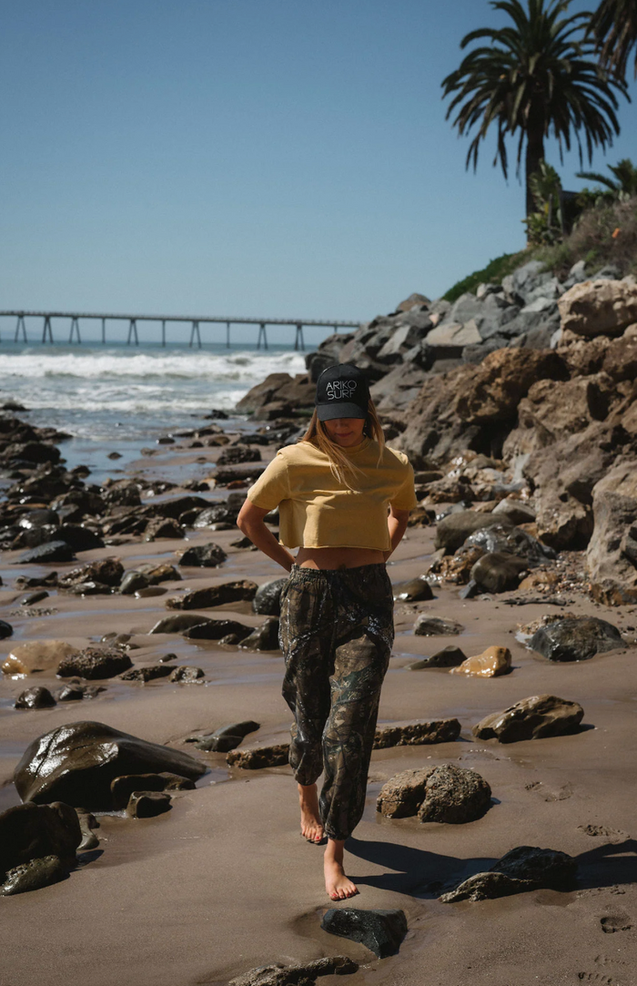 Person in yellow shirt and camouflage pants walking on a rocky beach with a bridge and palm trees in the background.