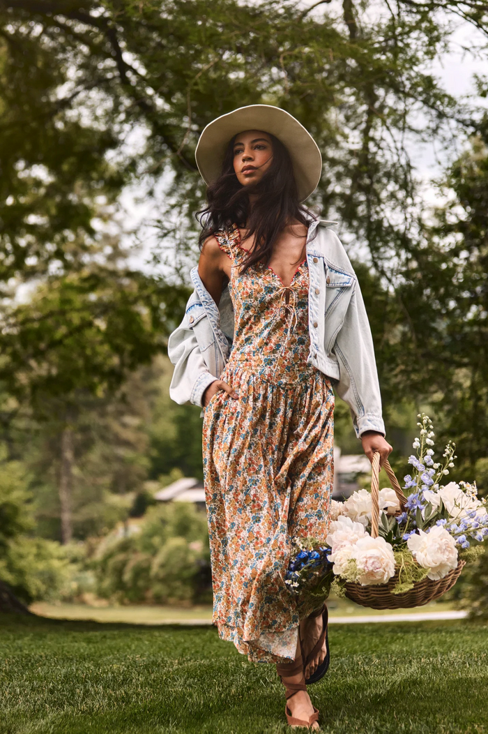 A woman walking outdoors on a grassy lawn wearing a bohemian-style floral maxi dress, a light blue denim jacket draped off her shoulders, and a wide-brimmed straw hat, while carrying a basket of white and purple flowers.