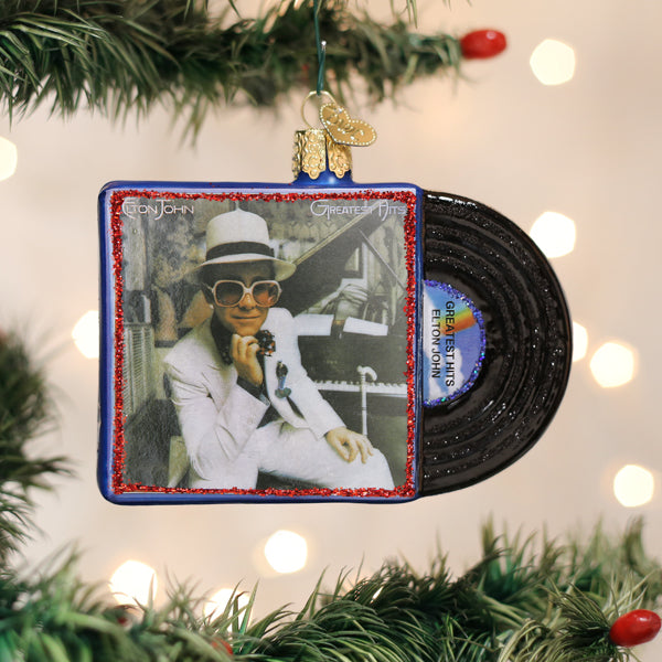 A Christmas tree ornament shaped like an Elton John vinyl record album hangs on a tree. The ornament features Elton John in a white suit and sunglasses, with a black record partially sticking out of the album cover.