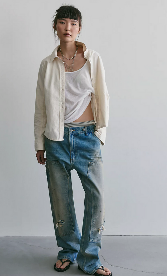 A person wearing distressed light blue wide-leg denim jeans with a faded wash, styled with an off-white tank top, an open cream shirt, and black sandals against a plain light gray wall.