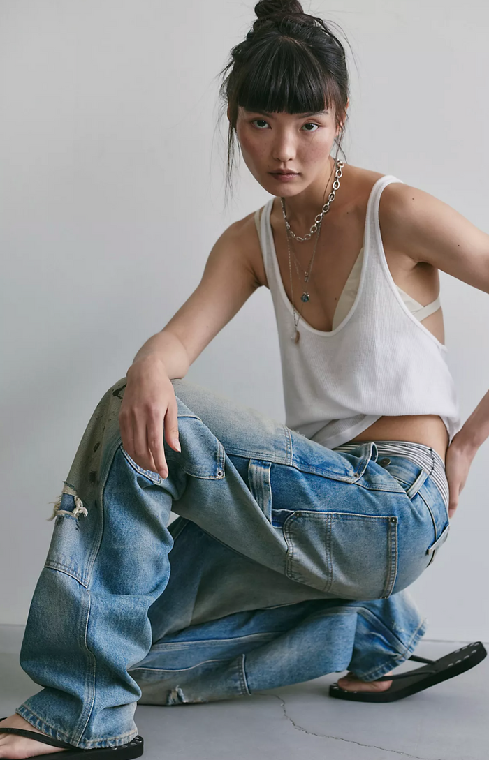 A model wearing a white ribbed tank top and distressed light blue wide-leg jeans, sitting on the floor against a plain light gray wall.