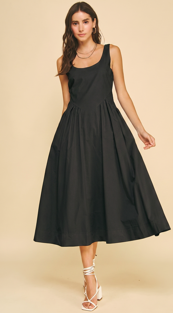 A woman modeling a sleeveless black midi dress with a scoop neckline, structured bodice, and full flared skirt, paired with white lace-up heels against a plain beige studio background.