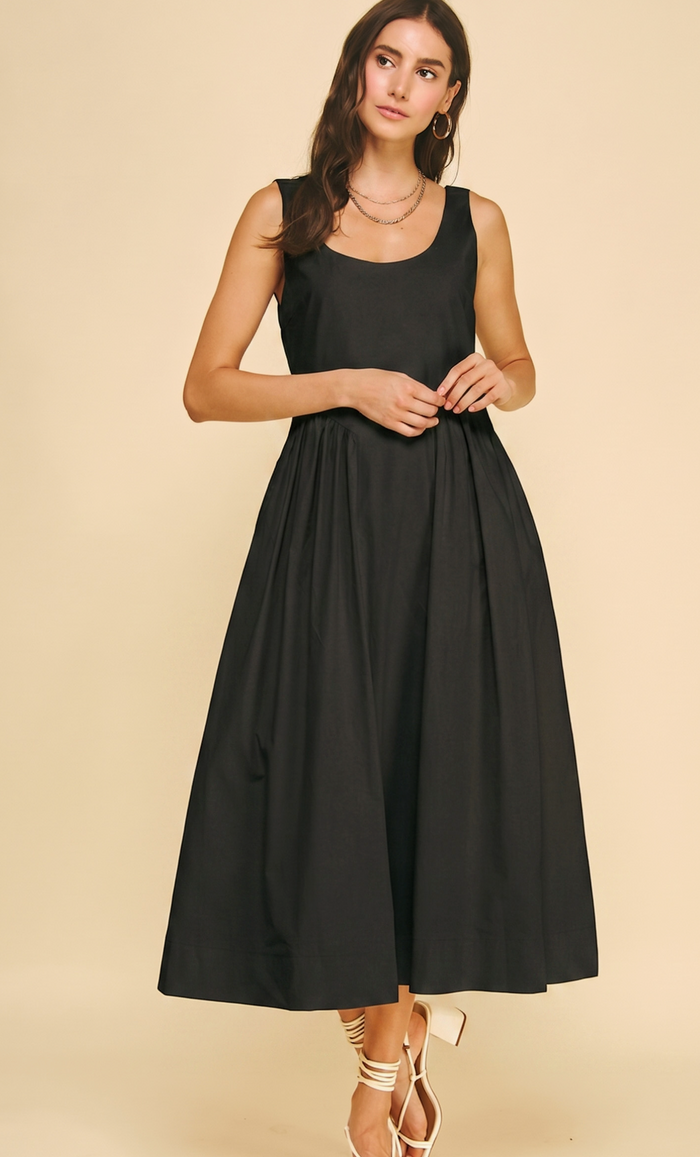 A woman modeling a sleeveless black midi dress with a scoop neckline and a flared skirt, standing against a solid beige studio background.