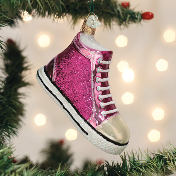 A pink glittery sneaker ornament hangs on a Christmas tree, with blurred white lights glowing softly in the background.