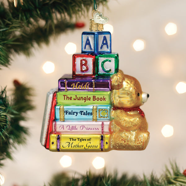 A Christmas tree ornament shaped like a gold teddy bear sitting next to a stack of colorful books and ABC blocks, hanging on a tree with white lights in the background.