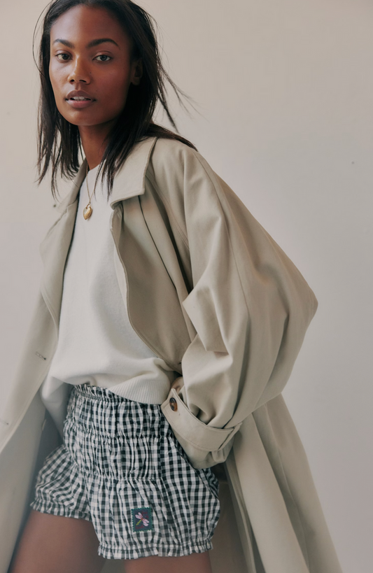 Woman wearing a beige trench coat, white shirt, and checkered shorts against a plain background