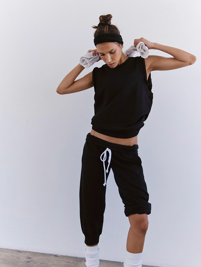 Woman in black sleeveless top and black jogger pants with white drawstring, standing against a white background.