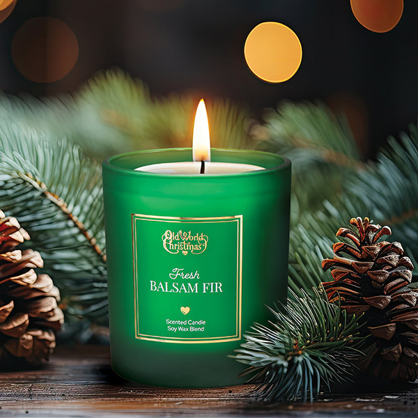 A lit green candle labeled Fresh Balsam Fir sits surrounded by pinecones and fir branches, with warm, blurred lights in the background, creating a festive holiday atmosphere.