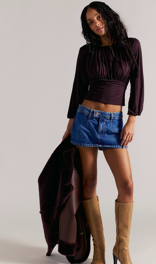 Woman wearing a dark brown top, blue denim skort and tan knee-high boots on a plain background