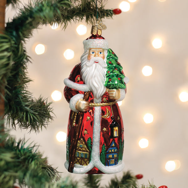 A glass Santa Claus ornament holding a green Christmas tree hangs on a decorated tree branch, with warm white lights blurred in the background.