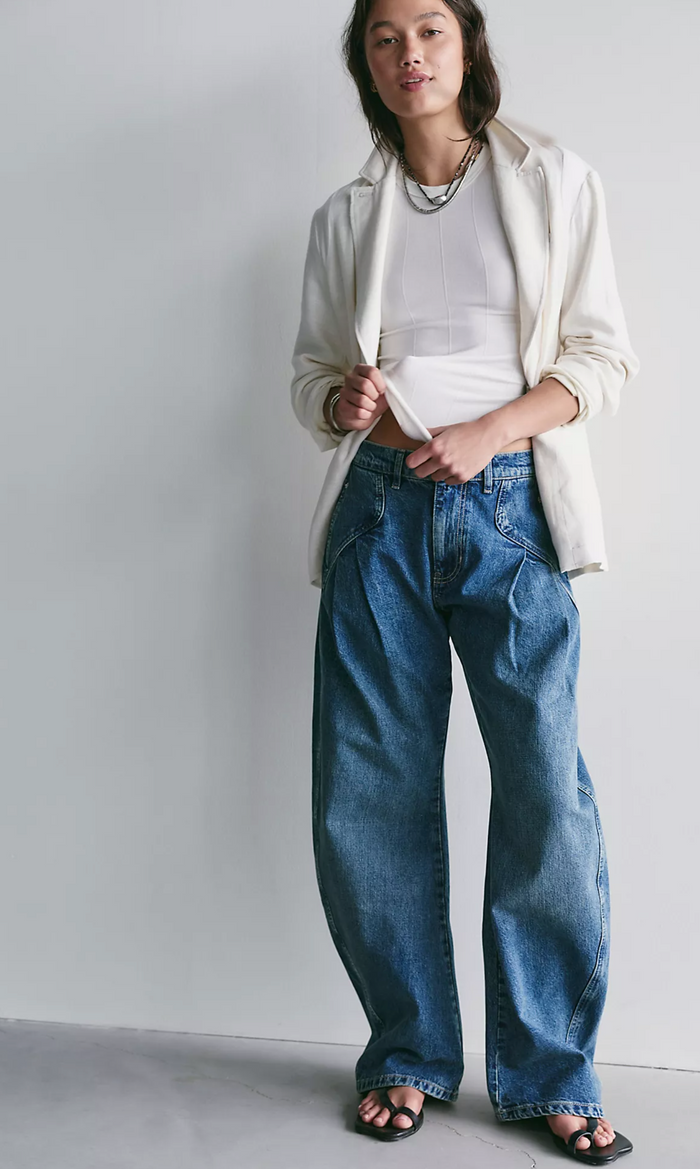 Person wearing a white jacket, white top, and blue jeans against a plain background