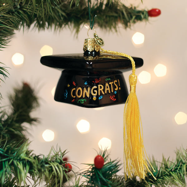 A black graduation cap ornament with a yellow tassel and the word CONGRATS! hangs on a Christmas tree branch, with festive lights softly glowing in the background.