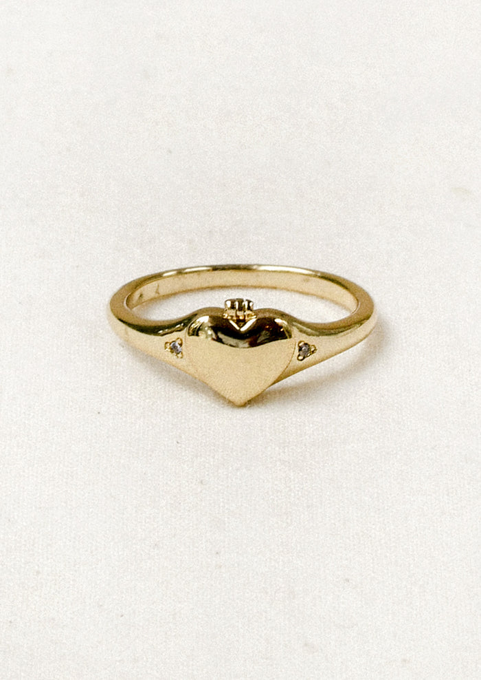 A gold ring with a tiny locket in the shape of a heart in the center and crystal detailing on either side.