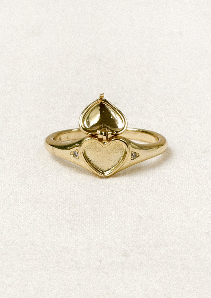 A gold ring with a tiny open locket in the shape of a heart in the center and crystal detailing on either side.