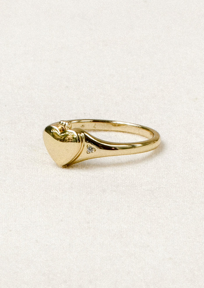 A gold ring with a tiny locket in the shape of a heart in the center and crystal detailing on either side.