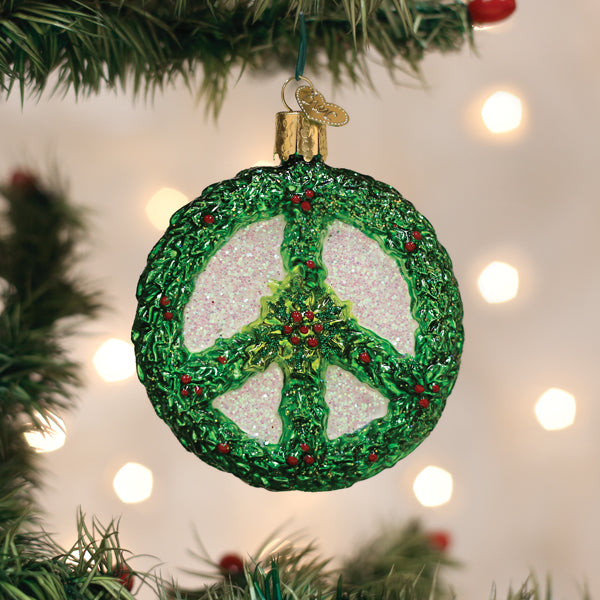 A Christmas tree ornament shaped like a green peace sign wreath with red berries hangs on a tree branch, with blurred white lights in the background.