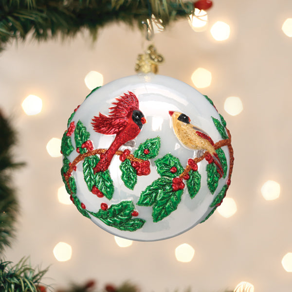 A white Christmas ornament decorated with red and yellow cardinals perched on holly branches with green leaves and red berries, hanging on a tree with twinkling lights in the background.