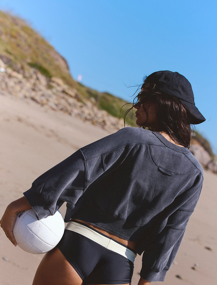 A woman on a sandy beach wearing a black cropped long-sleeve shirt, black swim trunks with a white waistband, and a black bucket hat, holding a white volleyball.